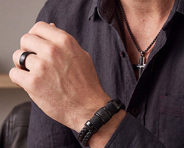A close-up of a man wearing a black beaded bracelet on his wrist and a black ring on his finger, wearing a dark button-up shirt and a black necklace with a silver cross pendant. The text 'MEN'S JEWELLERY' is displayed at the bottom.