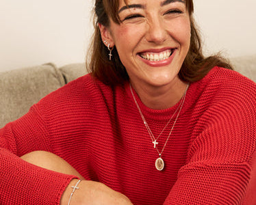A smiling woman wearing layered silver religious necklaces and a bracelet, with a red knit sweater, sitting on a couch. The text 'RELIGIOUS JEWELLERY' is overlaid at the bottom.