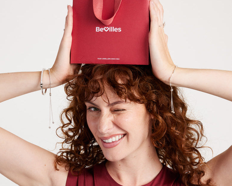 A smiling woman with curly brown hair holds a red BeOilles shopping bag above her head against a light gray background, wearing a matching burgundy top with the words 'GIFT GUIDE' overlaid at the bottom.