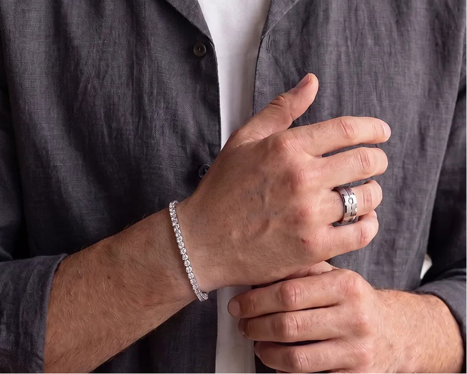 A close-up of a man wearing a silver tennis bracelet on his wrist and a silver ring on his finger, dressed in a dark gray linen shirt over a white t-shirt.