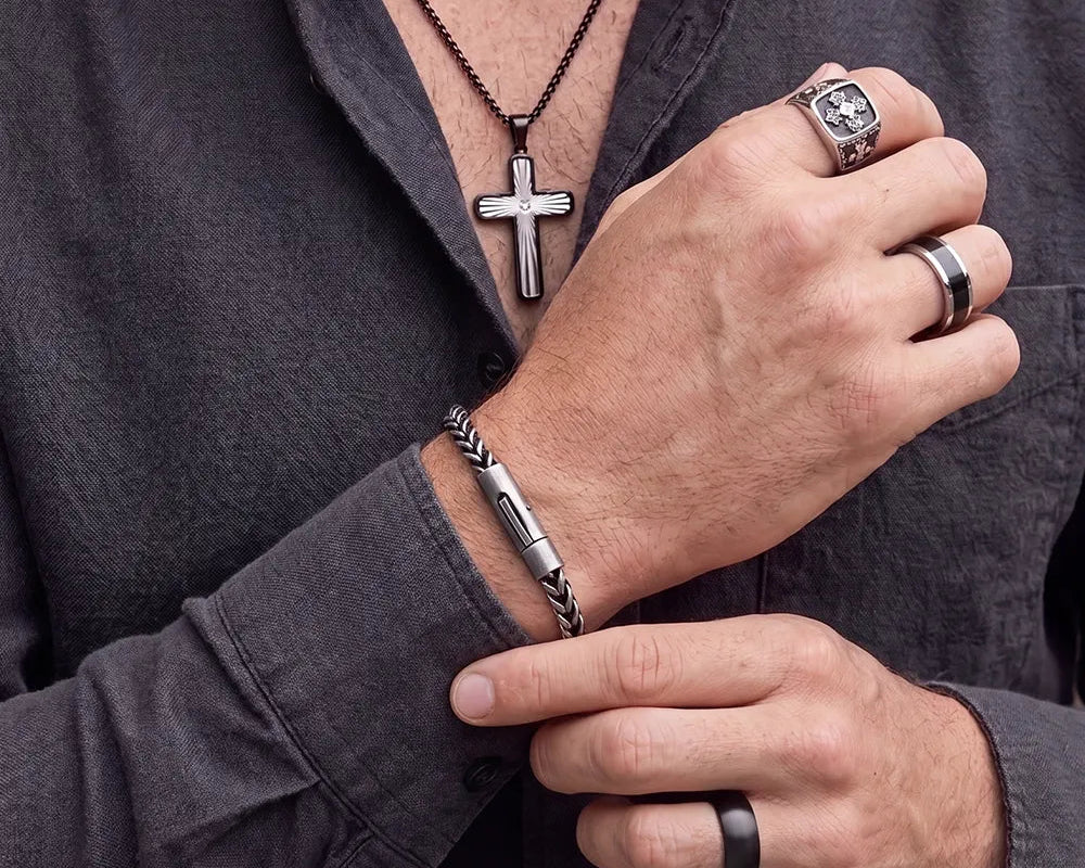 A close-up of a man's hands and chest wearing multiple silver-toned jewelry pieces, including a cross pendant necklace, a bracelet, a ring with a square face, and a band ring, over a dark gray button-down shirt.
