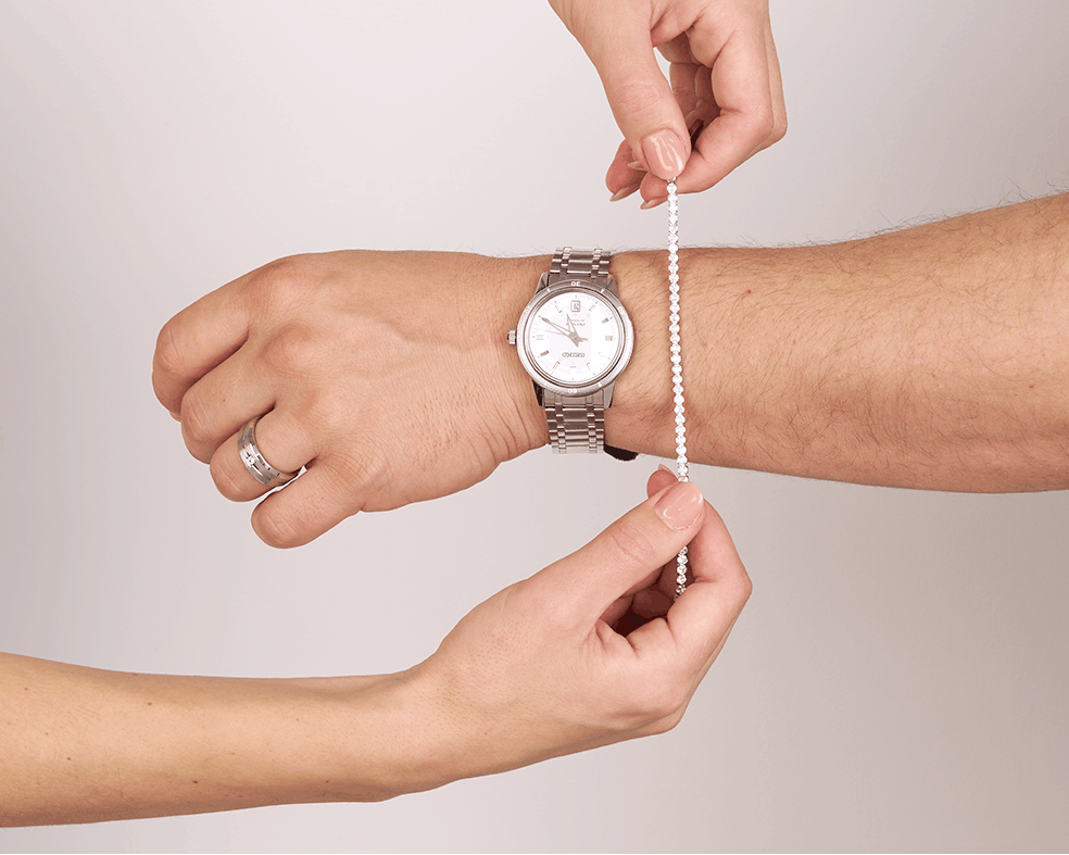 A close-up of a person's wrist wearing a silver metal watch, with two hands holding a thin white beaded measuring chain against the wrist to show the watch band size, on a light gray background.