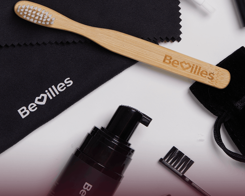 A bamboo toothbrush engraved with the Be\u00e9villes logo lies on a black microfiber cloth, with additional Be\u00e9villes-branded skincare accessories arranged around it on a white surface.