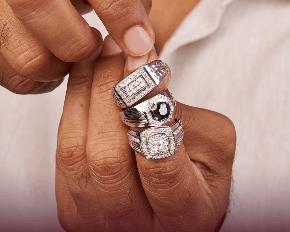 A close-up of a person's hand wearing three silver-toned rings with square-cut stones, including a black gemstone accent in the center, against a white shirt background.