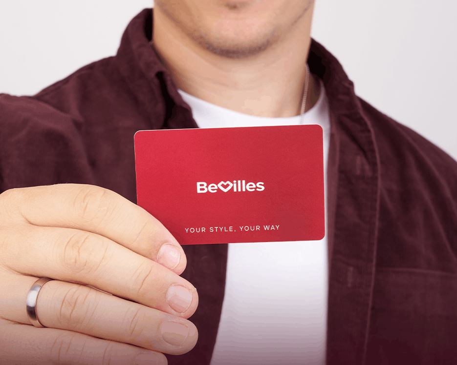 A smiling man in a maroon shirt holds up a red BeVilles gift card in front of his face against a light gray background.