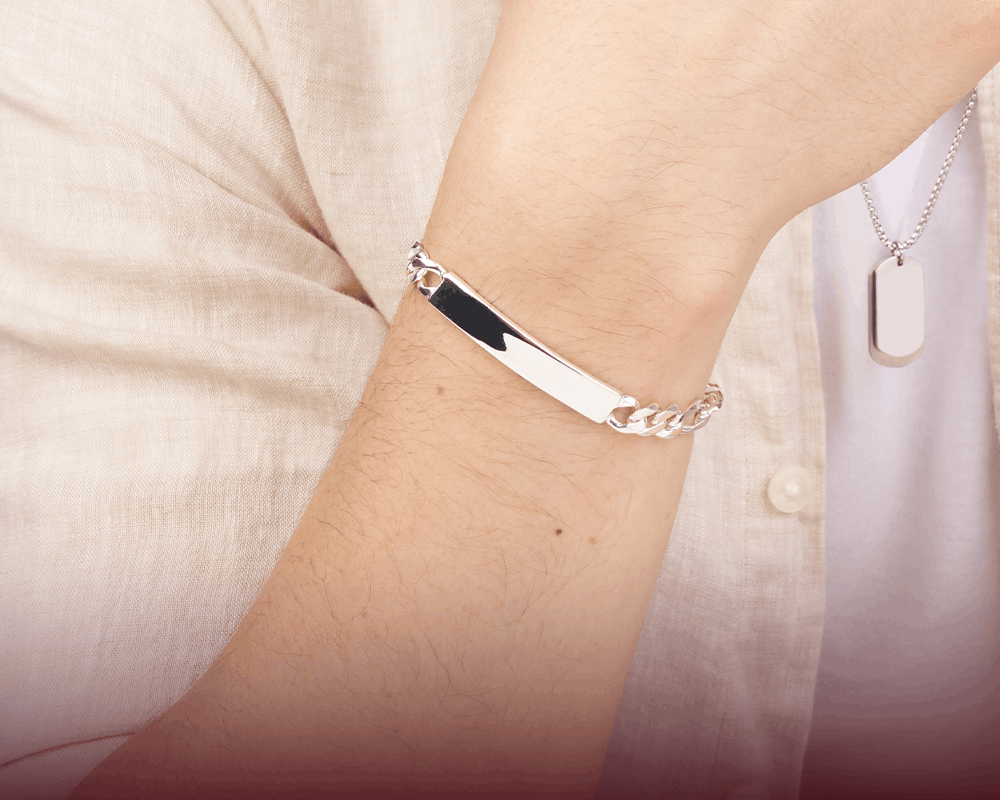 A polished silver ID bracelet with a rectangular plate engraved with initials, worn on a person's wrist. The person is wearing a light beige linen shirt and a silver dog tag necklace.