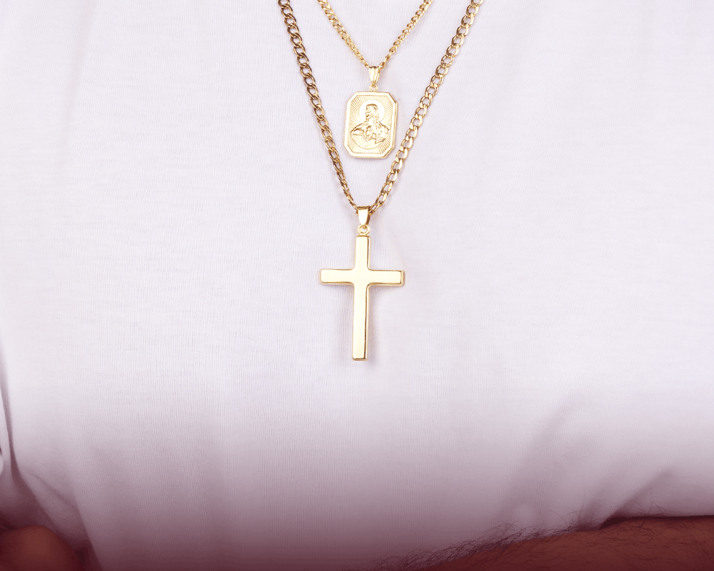 A close-up of a person wearing a layered gold necklace set. The shorter chain features a small oval religious pendant, while the longer chain holds a larger cross pendant.