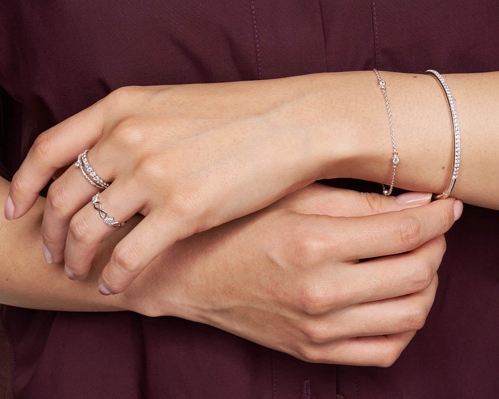 A close-up of a person's hands wearing delicate silver jewelry, including a thin silver chain bracelet on the wrist, a silver ring with a small heart, and a silver band with pavé stones, set against a dark burgundy button-up shirt.