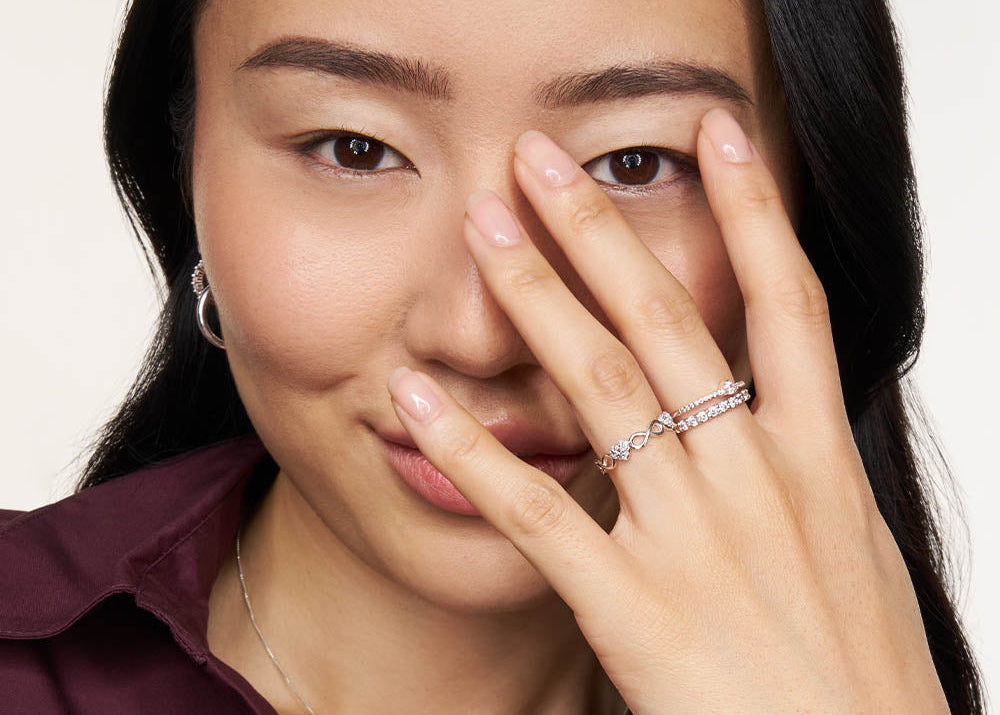 A close-up lifestyle shot of a woman wearing a delicate silver ring with small clear stones on one finger, holding her hand near her face against a clean white background.