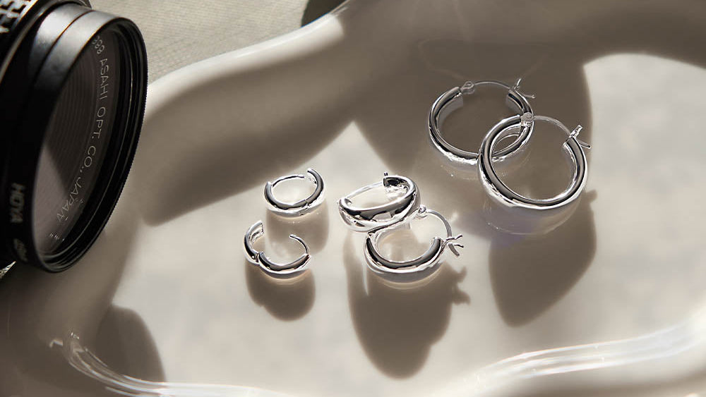 A set of polished silver hoop earrings displayed on a white wavy ceramic dish, styled with a camera lens and a small mirror in warm, directional sunlight.