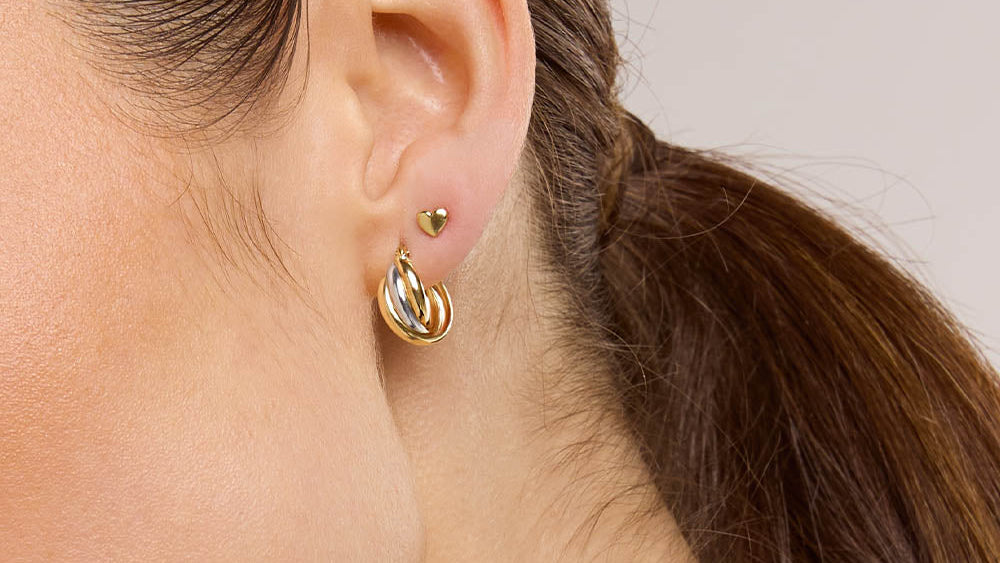 A close-up of a woman's ear wearing a pair of gold hoop earrings, with her hair tied back in a ponytail against a plain light background.