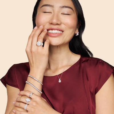 A woman with closed eyes and a smile wears multiple silver-toned jewelry pieces, including a ring on her finger, a necklace, bracelets, and earrings, against a solid beige background.