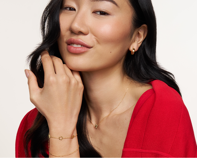 A close-up lifestyle shot of a woman with long black hair wearing delicate gold jewelry, including a small gold hoop earring, a delicate gold necklace, and a thin gold bracelet, paired with a vibrant red top against a clean off-white background.