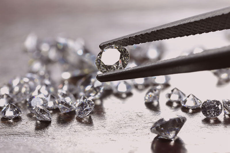 Diamonds being held by tweezers on a wooden surface with other diamonds scattered around.